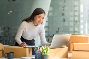Happy young Asian business owner woman prepare parcel box and check online orders of product for deliver to customer on laptop computer. Shopping Online concept