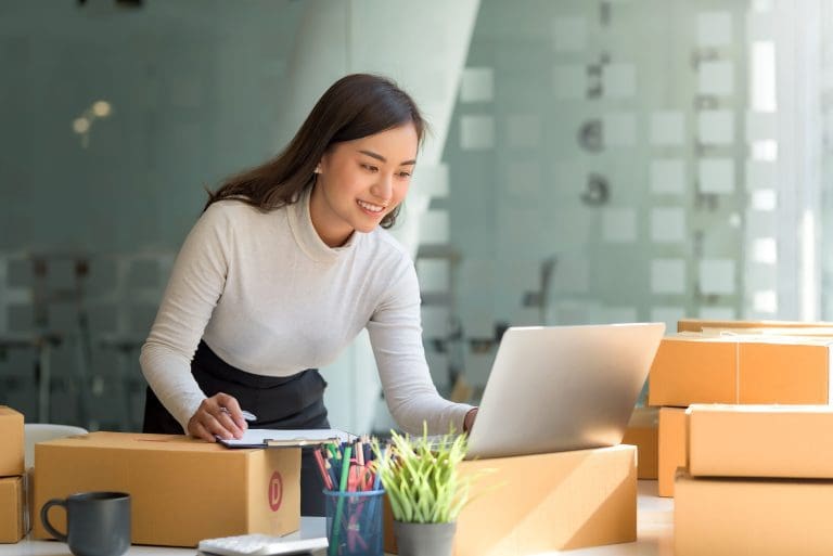 Happy young Asian business owner woman prepare parcel box and check online orders of product for deliver to customer on laptop computer. Shopping Online concept