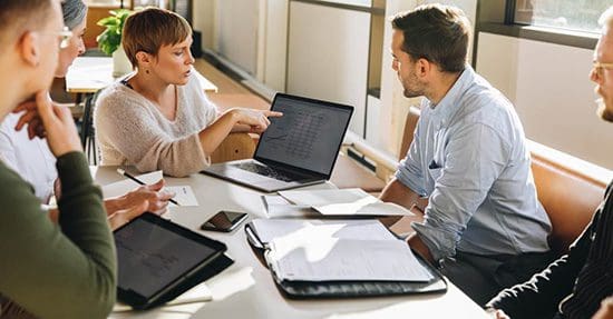 Manager pointing at laptop screen and talking to colleagues during business meeting. Female executive showing data to team in meeting.