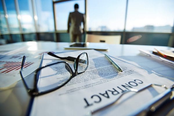 Man standing in front of office window while his glasses lay on his business documents on the table