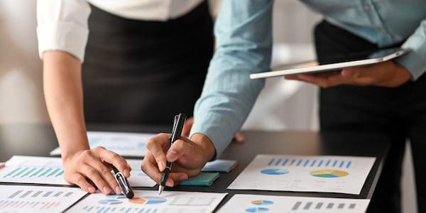 Cropped image of data analyst team working together with computer tablet and graphic charts information while standing together at modern meeting desk over meeting room as background.